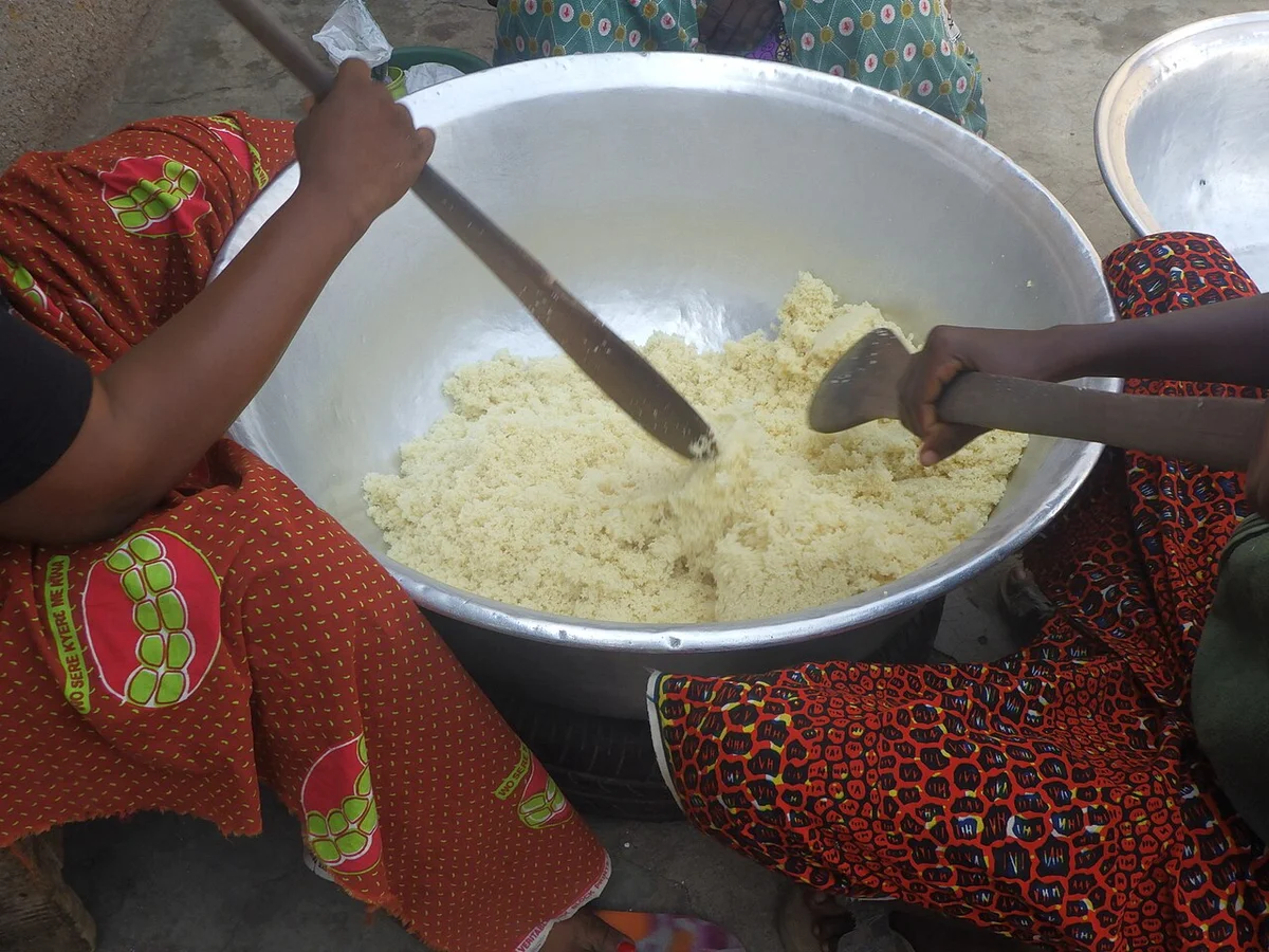 大きな鍋でアチェケを蒸している工程。複数の女性が木べらで混ぜている